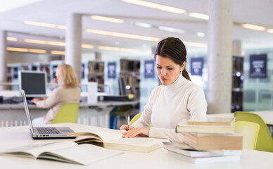Thoughtful woman working with textbook and laptop in library. High quality photo