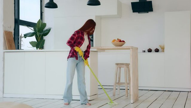 Young neat housewife redhead woman with long hair girl from cleanup agency take care of home house cleanliness washes floor with mop cleaning apartment modern kitchen room wipes wet scrubbing parquet