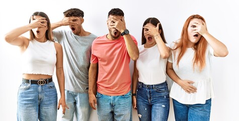 Group of young friends standing together over isolated background peeking in shock covering face and eyes with hand, looking through fingers with embarrassed expression.