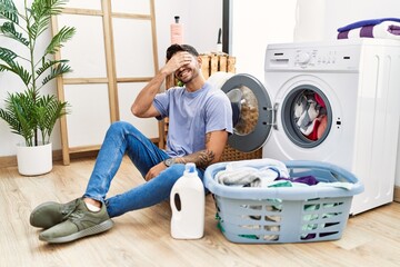 Young hispanic man putting dirty laundry into washing machine smiling and laughing with hand on face covering eyes for surprise. blind concept.