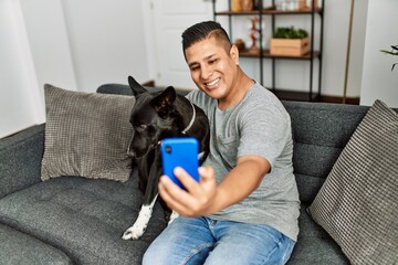 Young hispanic man make selfie by the smartphone sitting on the sofa with dog at home.
