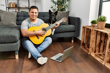 Young hispanic man having onlinr classical guitar lesson sitting on the floor with dog at home.