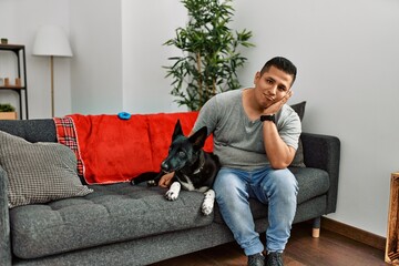 Young latin man and dog sitting on the sofa at home thinking looking tired and bored with...