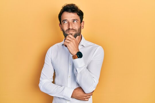Handsome man with beard wearing casual white t shirt thinking concentrated about doubt with finger on chin and looking up wondering