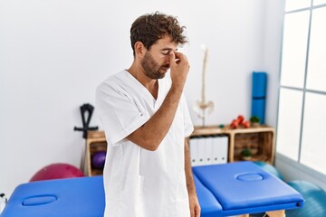 Young handsome physiotherapist man working at pain recovery clinic tired rubbing nose and eyes feeling fatigue and headache. stress and frustration concept.