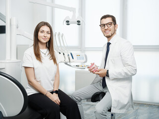 Fototapeta premium Happy male dentist sitting near dental unit with caucasian female patient with a smile looking at camera