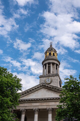 Fototapeta na wymiar London, Great Britain - July 3, 2022: Closeup of clock tower with golden caryatid statues and pediment on columns of St. Marylebone Parish Church under blue cloudscape. Green foliage 