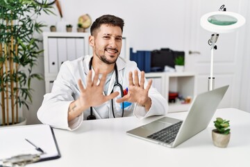 Young doctor working at the clinic using computer laptop afraid and terrified with fear expression stop gesture with hands, shouting in shock. panic concept.