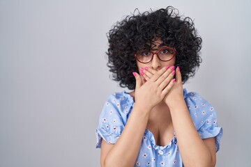 Young brunette woman with curly hair wearing glasses over isolated background shocked covering mouth with hands for mistake. secret concept.