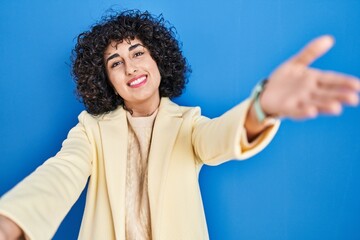 Fototapeta na wymiar Young brunette woman with curly hair standing over blue background looking at the camera smiling with open arms for hug. cheerful expression embracing happiness.
