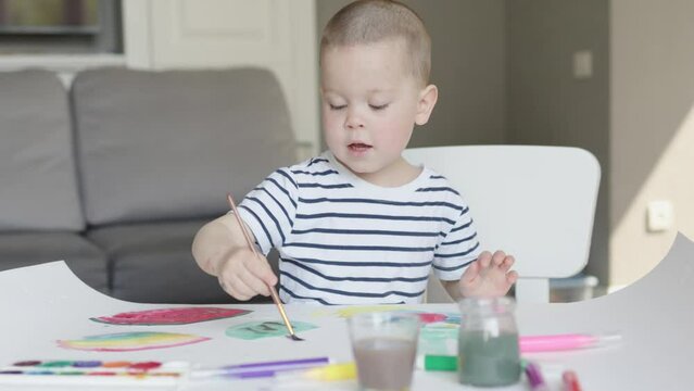 happy kid boy child art painting, talking, sitting at table home in living room. cute enthusiastic caucasian child drawing colorful picture artwork using paintbrush and watercolor aquarelle paints