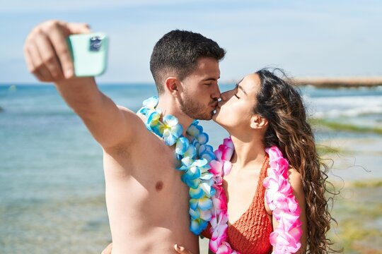 Young hispanic couple tourists wearing hawaiian lei make selfie by smartphone at seaside