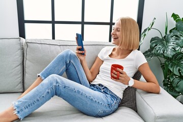 Young blonde woman drinking coffee using smartphone at home