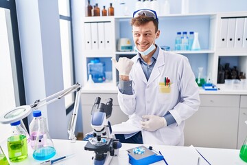Caucasian man working at scientist laboratory pointing thumb up to the side smiling happy with open mouth