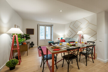 Living room with wooden dining table with black chairs and crockery with trays of fruit and glasses with orange juice, balcony with bay window and eclectic decoration and lots of artificial plants