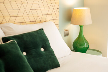 Corner of a bedroom with a headboard with pillows, green cushions and a lamp with a green porcelain base