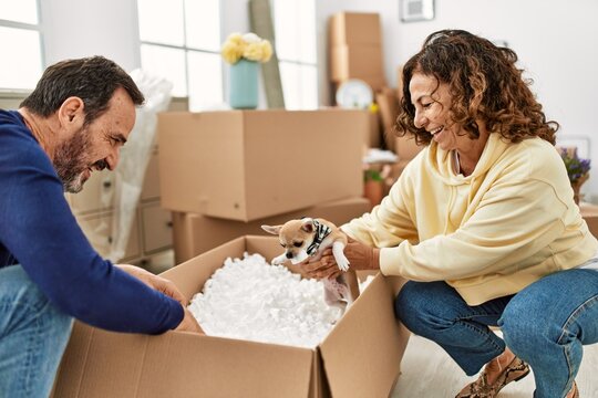 Middle age hispanic couple smiling happy playing with dog and foam package at new home.