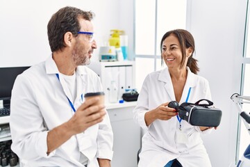 Obraz premium Middle age man and woman partners wearing scientist uniform drinking coffee and holding vr goggles at laboratory