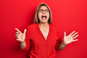 Middle age hispanic woman wearing casual clothes and glasses crazy and mad shouting and yelling with aggressive expression and arms raised. frustration concept.