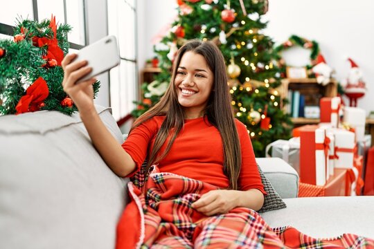 Young latin woman make selfie by the smartphone sitting by christmas tree at home