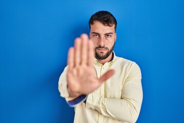 Handsome hispanic man standing over blue background doing stop sing with palm of the hand. warning...