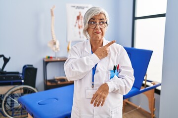 Middle age woman with grey hair working at pain recovery clinic pointing aside worried and nervous with forefinger, concerned and surprised expression