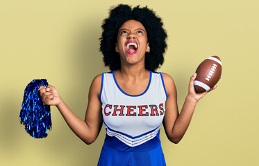 Young african american woman wearing cheerleader uniform holding pompom and football ball angry and...