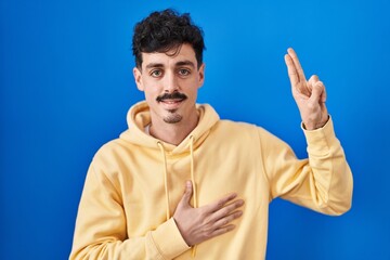 Naklejka premium Hispanic man standing over blue background smiling swearing with hand on chest and fingers up, making a loyalty promise oath
