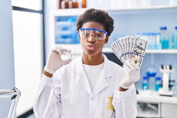 African american woman working at scientist laboratory holding dollars looking at the camera blowing a kiss being lovely and sexy. love expression.
