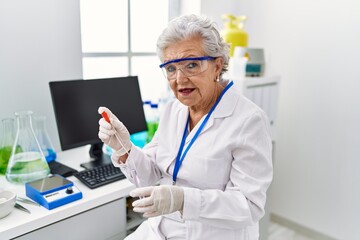 Senior grey-haired woman wearing scientist uniform using pipette and test tube at laboratory