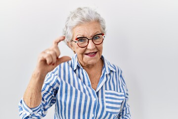 Senior woman with grey hair standing over white background smiling and confident gesturing with hand doing small size sign with fingers looking and the camera. measure concept.