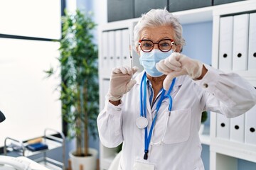 Senior woman with grey hair wearing doctor uniform and medical mask holding syringe with angry...