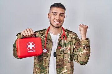 Young hispanic doctor wearing camouflage army uniform holding first aid kit screaming proud,...