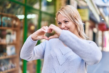 Young blonde woman smiling confident doing heart gesture with hands at street