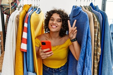Young hispanic woman searching clothes on clothing rack using smartphone smiling looking to the camera showing fingers doing victory sign. number two.