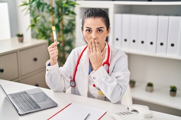 Young doctor woman holding blood sample covering mouth with hand, shocked and afraid for mistake....