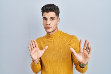 Young hispanic man standing over blue background moving away hands palms showing refusal and denial...