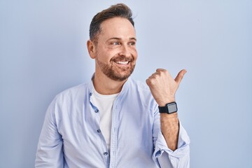 Middle age caucasian man standing over blue background smiling with happy face looking and pointing to the side with thumb up.