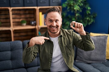 Middle age caucasian man holding virtual currency bitcoin pointing finger to one self smiling happy and proud