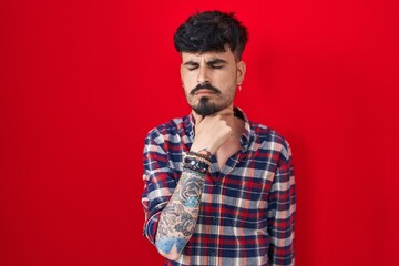 Young hispanic man with beard standing over red background touching painful neck, sore throat for...
