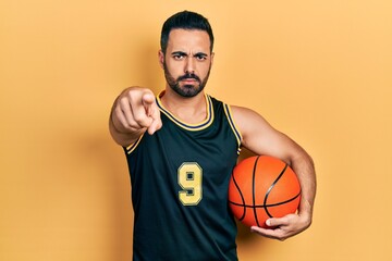 Handsome hispanic man with beard holding basketball ball pointing with finger to the camera and to...