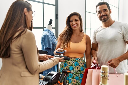 Young hispanic couple smiling happy paying using smartphone at clothes store.