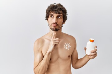 Young hispanic man standing shirtless holding sunscreen lotion thinking concentrated about doubt...