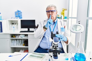 Senior caucasian man working at scientist laboratory asking to be quiet with finger on lips....