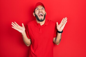 Hispanic man with beard wearing delivery uniform and cap celebrating mad and crazy for success with...