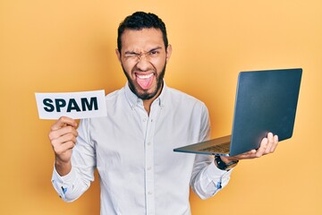 Hispanic man with beard working using computer laptop holding spam banner sticking tongue out happy...