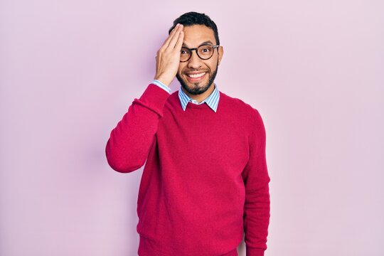 Hispanic man with beard wearing business shirt and glasses covering one eye with hand, confident smile on face and surprise emotion.