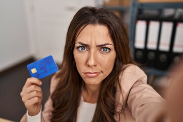 Young brunette woman working at small business ecommerce holding credit card skeptic and nervous, frowning upset because of problem. negative person.