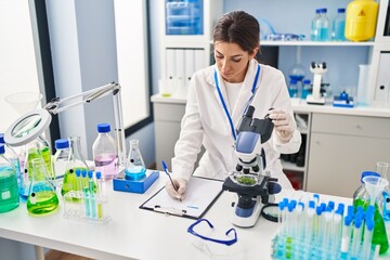 Young hispanic woman wearing scientist uniform using microscope and writing on clipboard at laboratory