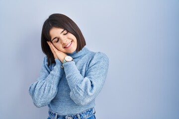 Young hispanic woman standing over blue background sleeping tired dreaming and posing with hands together while smiling with closed eyes.